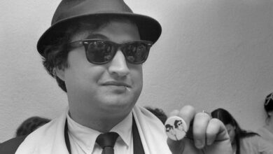 john belushi in suit and glassed holding blues brothers pin